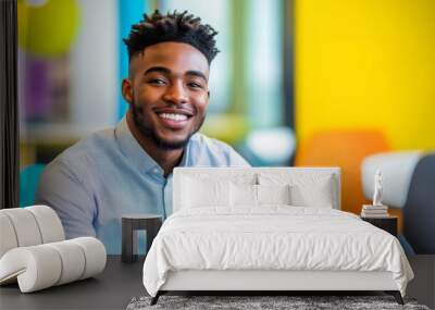 A young African American man exudes confidence as he sits in a bright, modern office. His cheerful smile reflects a positive work environment filled with color and energy Wall mural