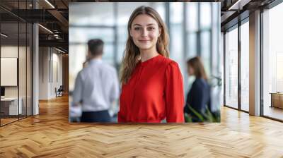 A young woman dressed in a vibrant red blouse smiles confidently in a contemporary office filled with colleagues engaged in work-related activities Wall mural