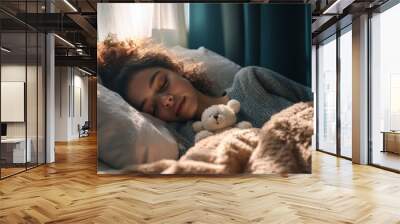 A young woman enjoys a peaceful sleep nestled in a cozy bed filled with soft pillows, clutching a teddy bear, as morning light gently filters through the curtains Wall mural