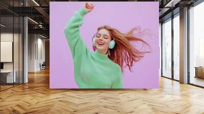 A young woman in a green sweater dances joyfully, wearing headphones, with her hair flowing, creating a lively vibe in a vibrant environment Wall mural