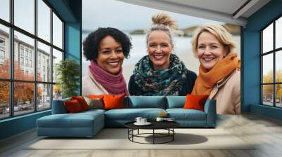 Three women of diverse backgrounds stand together on a beach, dressed in stylish clothing and warm accessories, sharing smiles on a cool day by the ocean Wall mural