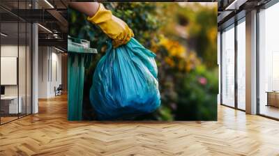 Wearing bright yellow gloves, an individual holds a blue trash bag, preparing to place it into a green trash bin against a backdrop of foliage in the evening light Wall mural