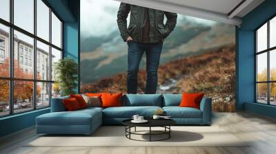 A man in a green jacket and jeans stands on a path in the mountains. Wall mural