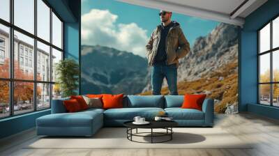 Young man in a brown jacket stands on a mountain path with scenic views. Wall mural