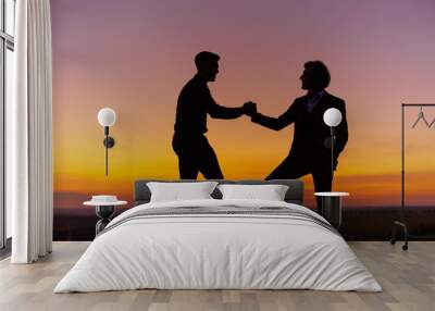 Silhouette of two men in sunset sky (orange and pink sky) on rock hill. Two gentlemen are taking, looking and agree each other, then shake hands. Agreement and cooperation concept Wall mural