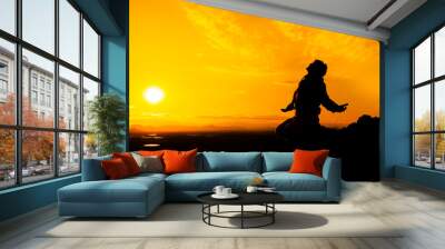 Silhouette of a person kneeling with open arms and looking at the sky on top of a mountain. Concept of religious and spiritual life. Warm atmosphere at dawn. Copy space on the right Wall mural