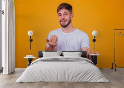 young handsome caucasian man wearing white t-shirt against yellow background inviting to come with hand has a warm smile and friendly expression on his face. happy that you came Wall mural