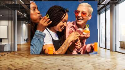 This is how we bond. Cropped shot of three attractive young women sitting against a blue wall together and bonding by blowing bubbles. Wall mural