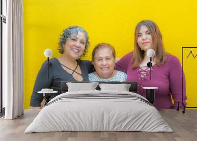 Daughter, grandmother and granddaughter hugging looking at the camera, three generations of Mexican women smiling in casual clothes on a yellow background Wall mural