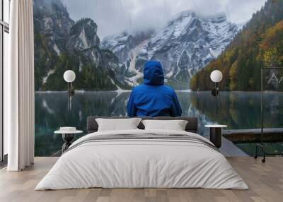 Young hiker in a blue jacket sitting on a wooden bridge beside a mountain lake gazing into the distance Wall mural