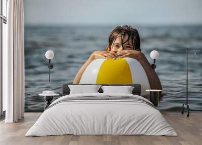 A boy holds a colorful beach ball while in the water, looking towards the camera. The background shows a calm sea under a sunny sky. Wall mural