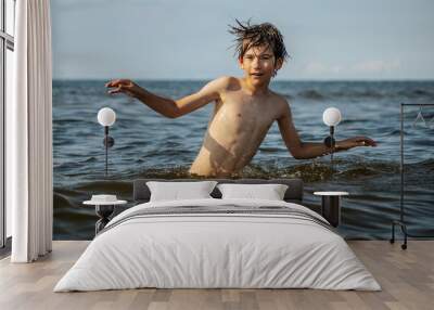 A boy joyfully splashes in the water with arms raised, capturing the excitement of playing in the ocean on a sunny day. The scene embodies the carefree spirit of childhood and summer fun Wall mural