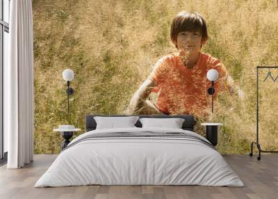 A young boy in an orange shirt sits cross-legged in a sunny meadow, meditating with hands in prayer position above his head. The scene captures a serene moment of mindfulness in nature. Wall mural