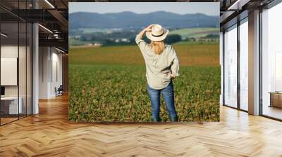 Woman with straw hat standing in agricultural field. Satisfied female farmer is looking at corn field in cultivated land Wall mural