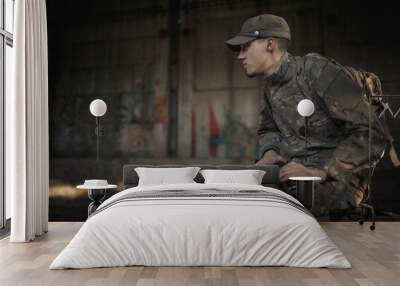 a soldier with a machine gun in a gray camouflage uniform wearing a cap and tactical black glasses sits on a concrete floor inside a large destroyed building Wall mural