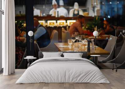 empty served table in an expensive restaurant for four people with glasses, plates and appliances in the background visitors and 3 cooks in white. evening interior of a popular cafe Wall mural