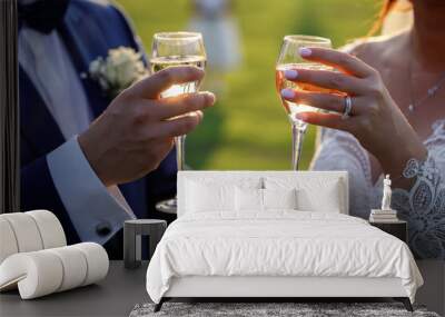 The groom in a blue classic suit and the bride in a white wedding dress, bow tie, with beautiful cufflinks, a large ring, drink champagne from glasses at sunset. Hands without faces close-up. Wall mural