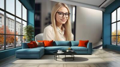 A young woman with glasses smiles while working at a computer desk in a bright, modern office setting.. Wall mural