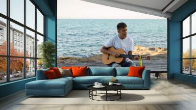 A young stylish guy plays the guitar on the embankment by the sea, the concept of music and relaxation Wall mural