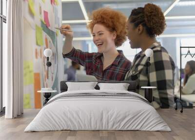 Two diverse female colleagues engage in a productive brainstorming session using sticky notes on a wall, embodying teamwork, creativity, and strategic planning in a modern office setting Wall mural