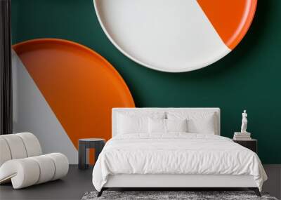 Two round plates with orange and white enamel are lying on a green background. One plate is placed higher than the other one Wall mural