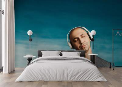 A bald man listens and enjoys music in large headphones with his eyes closed on a blue background. Wall mural