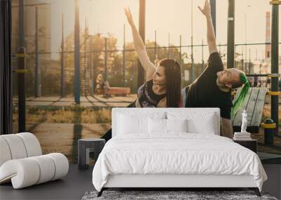 Young man and woman doing side plank synchronously with raised hand on the city sports ground. A girl with a hairstyle from many braids, a guy with an mohawk hairstyle Wall mural