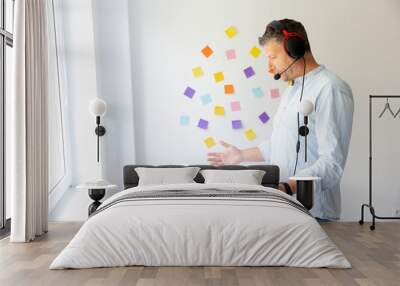 Man with headset stands at height adjustable table and works with notebook, behind him wall with colorful notes Wall mural