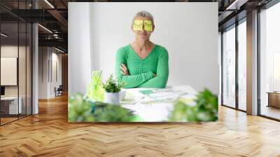 Middle-aged woman with short hair and green top is sitting at office table with many green plants and working notes and has sticky notes with painted eyes on her face Wall mural