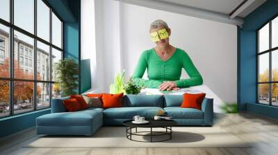 Middle-aged woman with short hair and green top is sitting at office table with many green plants and working notes and has sticky notes with painted eyes on her face Wall mural