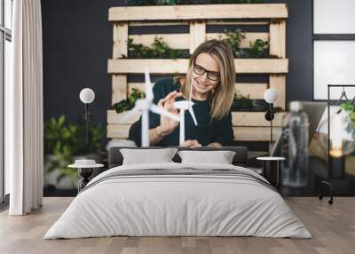 pretty, young and blond woman with stylish, modern black glasses sits in a sustainable office and works with pinwheel wind turbine models Wall mural