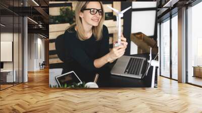 pretty, young and blond woman with stylish, modern black glasses sits in a sustainable office and works with pinwheel wind turbine models Wall mural