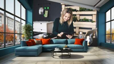 pretty young blond woman is sitting in a modern, sustainable office with many green ecological plants and is working on her tablet Wall mural