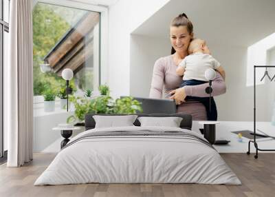 pretty young woman, businesswoman stands at work desk holding her sleeping son in her arms and working on notebook, smartphone and is happy Wall mural