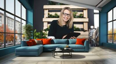 pretty young woman with glasses sits in a modern, sustainable office with lots of green ecological plants and works on her tablet and is happy and is smiling Wall mural