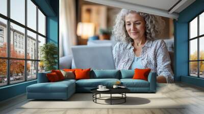 Happy curly middle aged woman using pc laptop surfing online sitting on sofa at home. Mature older lady looking away relaxing on couch with computer device in modern house living room Wall mural