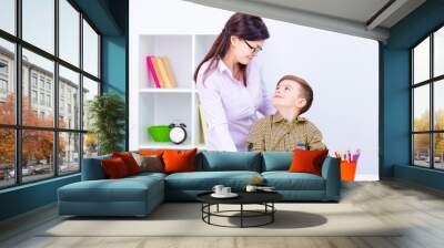 A boy sits at a table and writes in a notebook. A teacher with glasses stands next to him and looks at the boy. Wall mural