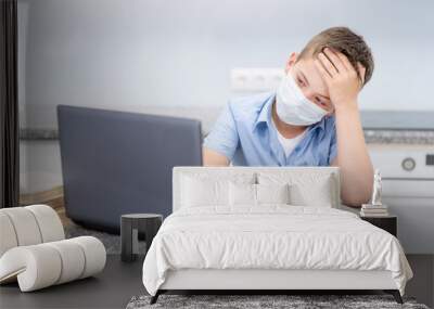 A boy with a blue shirt and a medical mask on his face is engaged in education using a laptop at home, he tiredly laid his head on his hand. Distance learning during sickness, online learning Wall mural