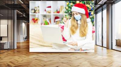 A young girl with a medical mask and a Santa hat holds a gift in her hands and makes a call online for Christmas (new year) to relatives. Coronavirus pandemic during the winter holidays. Lockdown at C Wall mural