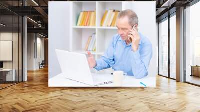 Senior talking on a cell phone near a laptop on the background of shelving with books. Hardware problems, work difficulties Wall mural