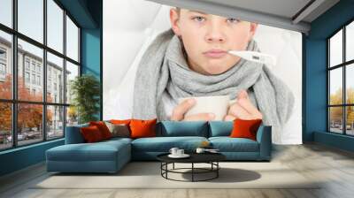 Sick boy sitting at home on the bed with a hot drink in his hands and measuring the temperature. Disease season Wall mural