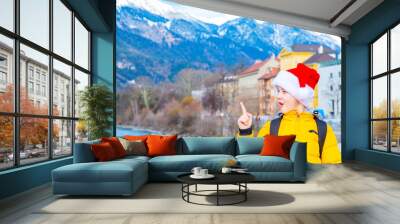 Smiling boy wearing red santa. The child points with an index finger to an empty place near him on the background of alpine mountains Wall mural