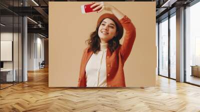 A young woman dressed in an orange jacket and brown hat takes a smiling selfie with a red phone against a beige background, capturing a joyful moment. Wall mural