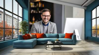 Portrait of handsome businessman smiling at camera while sitting in front of computer at the office desk Wall mural