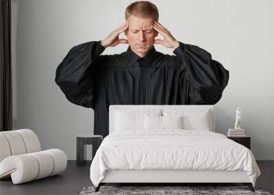 Caucasian male judge in a black robe, stressed and contemplative, with hands on his head against a neutral background. Wall mural