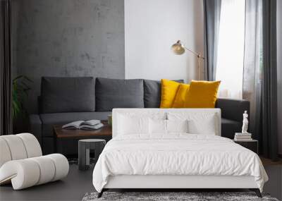 Gray sofa with yellow pillows in the living room.Green plant. Geometrical carpet. Book on the wooden table. Daylight. Empty gray concrete wal. Wall mural