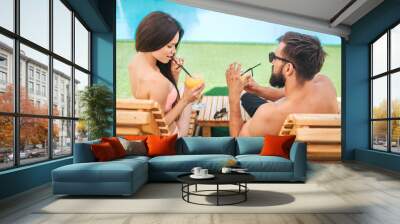 Attractive couple is sitting on sunbeds and drinking cocktails. Girl looks down. She holds straw. Guy wears glasses. Wall mural