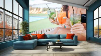 Cut view of three beautiful women sit on sunbeds and hold cocktails in hands. There are two green and one orange. Models cheer to relaxation and good rest. Wall mural