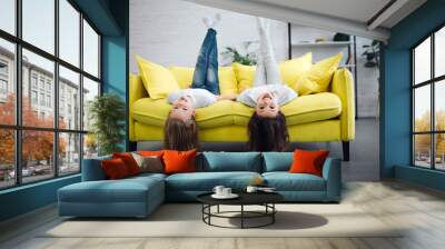 Funny young teenagers lying on yellow sofa and looking straigt on camera. They smile. Girls hold legs up in air. Their hair touch floor. Wall mural