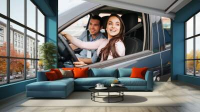 Male auto instructor takes exam in young woman. Cheerful attractive female driver posing at driver's place. Look from car. Hold hands on sheering wheel. Wall mural